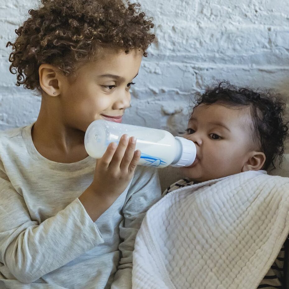 Content ethnic boy in domestic wear feeding adorable sibling with milk bottle while sitting together on comfy armchair